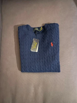 Mörkblå kabelstickad tröja Polo Ralph Lauren - Snygg mörkblå kabelstickad tröja från Polo Ralph Lauren med klassisk röd logga broderad på bröstet. Helt ny, priset kan diskuteras!