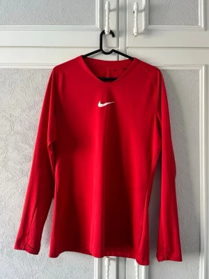Röd långärmad träningströja Nike - Snygg röd långärmad träningströja från Nike med vit logga på bröstet. Tröjan har rund halsringning och är tillverkad i ett mjukt, stretchigt material som passar perfekt för sport och träning.