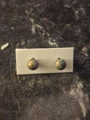 Små runda örhängen i metall - Säljer ett par små, runda örhängen i metall med en skimrande yta i guld och mörkgrönt. Örhängena har en strukturerad, nästan pärlliknande yta som ger dem en cool och unik look. Perfekta för dig som gillar subtila men ändå speciella accessoarer.