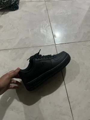 Svarta Nike Air Force 1 sneakers - Klassiska helsvarta Nike Air Force 1 sneakers i lågt utförande. Skorna är tillverkade i slätt läder med matchande svarta skosnören och sula. Ikonisk Swoosh på sidan och diskret Air-detalj på sulan. Perfekta för en clean och stilren look.