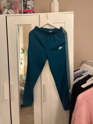 Blå mjukisbyxor från Nike - Säljer ett par gröna mjukisbyxor från Nike med vita ränder längs sidorna och Nike-logga på vänster ben. Byxorna har resår i midjan och är tillverkade i ett mjukt, sportigt material. Perfekta för chill eller träning.