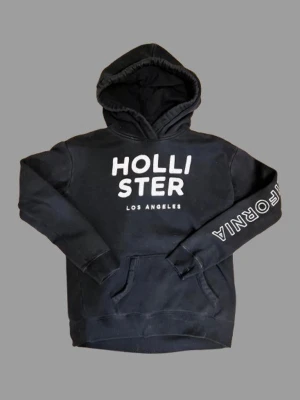 Svart hoodie från Hollister - Svart hoodie från Hollister med stor vit logga på bröstet och texten 'CALIFORNIA' på ena ärmen. Klassisk känguruficka framtill och mjuk huva. Perfekt för en avslappnad och cool stil. Materialet känns mjukt och bekvämt.