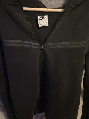 Svart Nike hoodie med dragkedja - Svart hoodie från Nike med hel dragkedja framtill och diskret Nike-logga på bröstet. Hoodien har en huva och tunna reflexdetaljer över bröstet. Tillverkad i mjukt material som känns skönt mot huden. Perfekt för en sportig och avslappnad stil. Den har en liten slitning som ni ser på bild 2