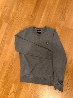 Grå crewneck från Lyle & Scott - En stilren grå sweatshirt från Lyle & Scott med rund halsringning och ribbade muddar. Tröjan har det klassiska gula örnlogot på bröstet. Tillverkad i mjukt material som känns skönt mot huden. Perfekt för dig som gillar enkel och clean stil. Dm:a gärna för fler bilder.🙂