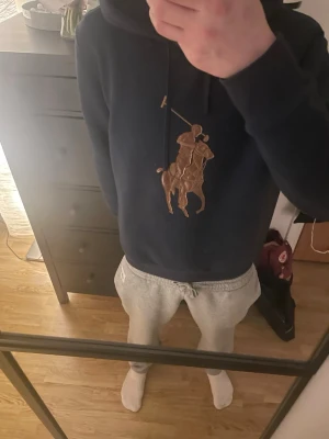 Mörkblå hoodie från Polo Ralph Lauren - Säljer en mörkblå hoodie från Polo Ralph Lauren med det klassiska stora polospelar-trycket i brunt och beige på bröstet. Hoodien har huva med dragsko och är tillverkad i mjukt material som känns skönt mot huden. Perfekt för en avslappnad och stilren look.