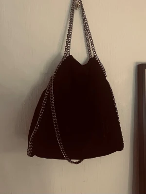 Svart kedjehandväska med axelrem - Snygg svart handväska med silverfärgade kedjedetaljer och axelrem. Väskan har en mjuk, slouchy form och är tillverkad i ett material som liknar mocka. Perfekt för dig som gillar en edgy och trendig stil