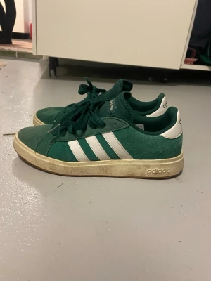 Gröna Adidas sneakers med vita ränder - Snygga gröna Adidas sneakers i mocka med klassiska vita ränder på sidorna och vit häl. Skorna har gröna skosnören och en beige sula med Adidas-logga. Perfekta för dig som gillar retrostil och streetwear.