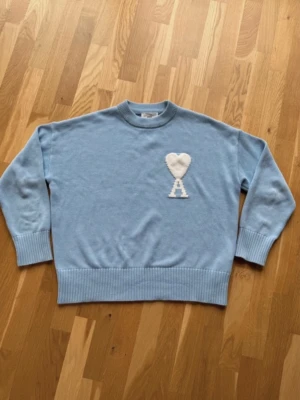 Ljusblå stickad tröja Ami Paris - Ljusblå stickad tröja från Ami Paris med rund halsringning och ribbade muddar. På bröstet finns ett vitt hjärta och bokstaven 'A' i intarsia. Tröjan har en avslappnad passform och är perfekt för lager på lager. Materialet känns mjukt och skönt.