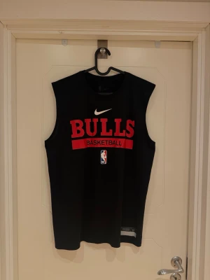 Svart Bulls basketlinne från Nike - Snyggt svart basketlinne från Nike med stort rött 'BULLS' tryck på bröstet och NBA-logga. Linnet är ärmlöst och tillverkat i Dri-Fit material som andas, perfekt för basket eller träning. Klassisk rund halsringning och officiell Chicago Bulls design.                          Nypris ca 600kr. Modellen är 185cm lång och väger 82kg.