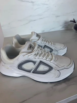  Dior B30 Sneakers - Snygga vita sneakers från Dior med grå och svarta detaljer. Skorna har en sportig siluett, meshpaneler och robust sula. Stora Dior-loggor på sidorna och klassisk snörning framtill. Perfekta för dig som vill ha en trendig och exklusiv look. (Har ingen låda till skorna)