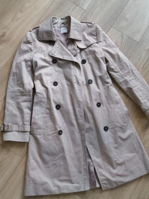 Beige trenchcoat med skärp - Stilren beige trenchcoat med dubbelknäppning, klassisk krage och bälte i midjan. Jackan har svarta knappar, axelklaffar och justerbara spännen vid ärmsluten. Tillverkad i ett lätt bomullsmaterial, perfekt för lager på lager. Fläckar enligt bild 4 och 5.