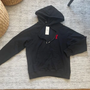 Svart hoodie från A.P.C. med röd detalj - Snygg svart hoodie från A.P.C. med klassisk huva och dragsko. På bröstet finns en liten röd broderad detalj som sticker ut. Hoodien har en stor magficka och ribbade muddar vid ärmslut och nederkant. Perfekt för en avslappnad och stilren look.