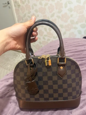 Louis Vuitton Damier Alma handväska - Snygg Louis Vuitton Alma handväska i klassiskt brunt Damier-mönster. Väskan har två stadiga handtag, guldfärgade metalldetaljer och ett hänglås framtill. Tillverkad i canvas med läderdetaljer och elegant rundad form.