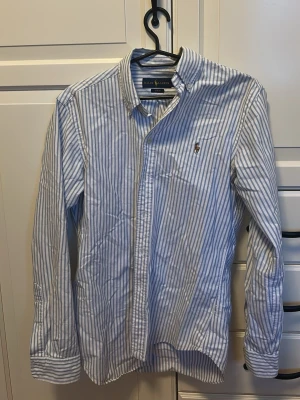 Blårandig skjorta från Ralph Lauren - Klassisk skjorta med blå och vita vertikala ränder från Ralph Lauren. Skjortan har lång ärm, knappslå framtill och en liten broderad logga på bröstet. Tillverkad i ett lätt bomullstyg och har en tidlös, avslappnad passform.