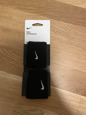 Svarta handledsband från Nike - Säljer ett par svarta handledsband från Nike med den klassiska vita swoosh-loggan broderad på framsidan. Perfekta för träning eller sport, tillverkade i mjukt och stretchigt material som absorberar svett och sitter bekvämt runt handleden.