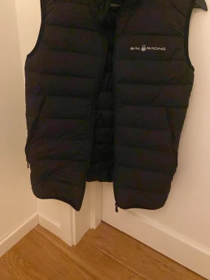 Svart dunväst från Sail Racing - Stilren svart dunväst från Sail Racing med diskret logga på bröstet. Västen har en klassisk quiltad design, hög krage och dragkedja framtill. Perfekt för lager-på-lager och håller dig varm under kyliga dagar. Materialet är lätt och smidigt, vilket gör den enkel att bära.