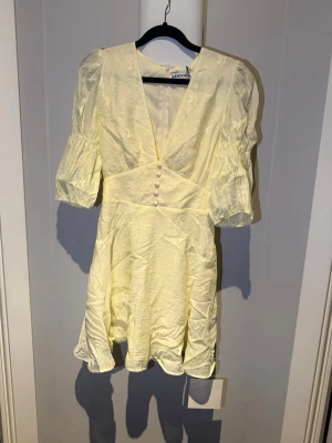 Ljusgul kort klänning från Adoore - Supersöt ljusgul kort klänning från Adoore med puffärmar, v-ringning, citroner och dekorativa knappar framtill. Aldrig använd med prislappen kvar.