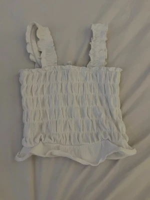 Vitt linne med volangband - Supersöt vit linnetopp med smockad struktur och breda volangprydda axelband. Toppen är ärmlös och har en lätt volangkant nertill. Perfekt för en fräsch och somrig look. Aldrig använt💛