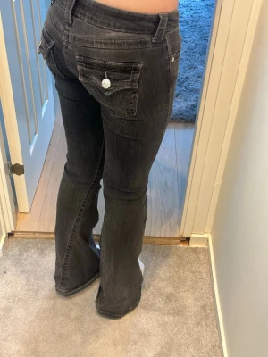 Svarta bootcut jeans True Religion - Säljer ett par svarta bootcut jeans från True Religion med snygga kontrastsömmar och stora fickor bak med vita knappar. Jeansen har låg midja och klassisk logga på bakfickan. Perfekta för dig som gillar en avslappnad men trendig stil. W25