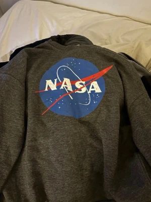 Grå NASA hoodie med tryck - Snygg grå hoodie med stort NASA-tryck på bröstet i blått, vitt och rött. Klassisk modell med huva och mjukt material, perfekt för dig som gillar rymdtema och vill ha en chill och trendig stil.
