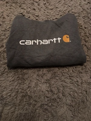 Mörkgrå Carhartt hoodie med logga - Snygg mörkgrå hoodie från Carhartt med stor vit logga och gul detalj på bröstet. Klassisk känguruficka framtill och huva med dragsko. Perfekt för dig som gillar streetwear och vill ha en bekväm och stilren look. Tillverkad i mjukt material. Det är en XS enligt lappen, men modellen är oversized så den ser större ut och passar snarare S/M beroende på hur man vill att den ska sitta🤗