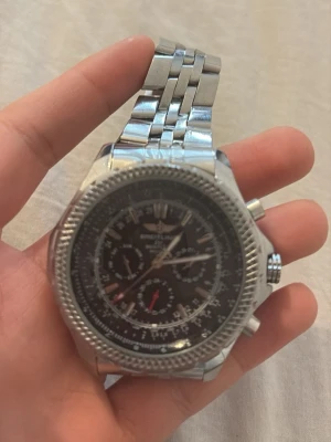 Silverfärgad Breitling kronografklocka - Stilren kronografklocka från Breitling med svart urtavla och flera subdials. Klockan har en robust länkad armlänk i silverfärgat metall och markerade detaljer runt boetten. Perfekt för dig som gillar klassisk och sportig design.