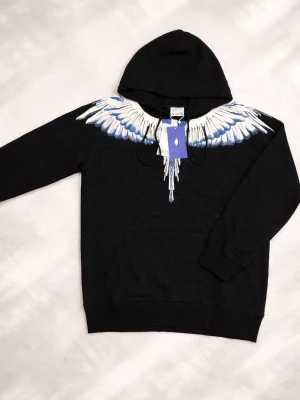 Marcelo burlon hoodie - Svart hoodie från Marcelo Burlon County of Milan med ikoniskt vingtryck i vitt och blått över axlar och bröst. Klassisk känguruficka framtill och justerbar huva. Perfekt för dig som vill sticka ut med en streetwear-look.