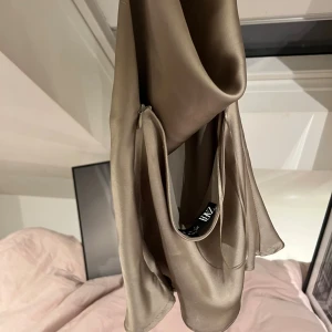 Champagnefärgad topp från ZARA - Säljer en champagnefärgad omlottopp från ZARA i glansigt satinliknande material i kashmir. Toppen har en stor rosett som knyts i ryggen och är ärmlös med elegant, flowy passform. Perfekt för dig som vill sticka ut med en chic och trendig look.