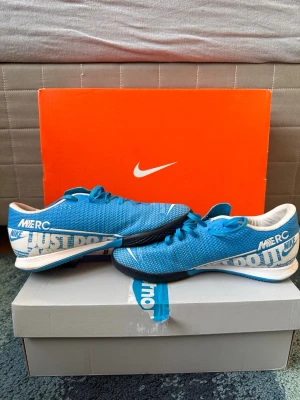 Nike Mercurial blå fotbollsskor - Säljer ett par Nike Mercurial fotbollsskor i blått med vita och svarta detaljer. Skorna har texten 'Just Do It' på sidan och är designade för bra grepp på planen. Lätta och smidiga med snörning och slitstark yttersula. Perfekta för dig som vill ha stil och prestanda på fotbollsplanen. Köp gärna med insta box så går de snabbare 