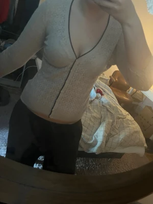 Beige figurnära cardigan med mönster - Snygg beige cardigan med diskret mönster och svarta kontrastsömmar längs knäppningen och halsringningen. Tröjan har djup urringning, långa ärmar och sitter tight mot kroppen. Perfekt för dig som gillar en stilren och trendig look.