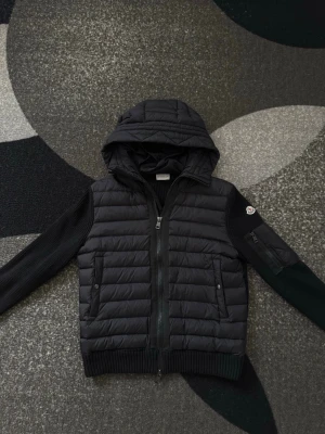Moncler cardigan - Snygg svart pufferjacka från Moncler med quiltad front och huva. Jackan har ribbstickade ärmar och muddar, två fickor med tryckknappar samt en ärmficka med Moncler-logga. Perfekt för kalla vinterdagar och riktigt trendig look. Knappt använd