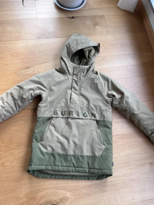 Beige och grön anorak från Burton - Säljer en snygg beige och grön anorak från Burton med stor huva och dragkedjor på fickorna. Jackan har BURTON-logga över bröstet och är tillverkad i slitstarkt material, perfekt för dig som gillar streetwear och outdoor-vibe.