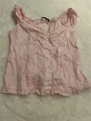 Rosa blus med volang från Brandy Melville - Söt ljusrosa blus från Brandy Melville med volangdetaljer vid axlar och nederkant. Blusen har knappar framtill, rynkade axelpartier och dekorativa veck. Tillverkad i mjuk bomull, perfekt för vår och sommar. 🥰