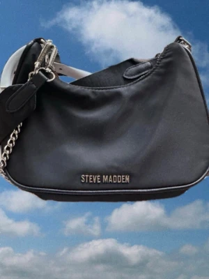 Svart axelremsväska Steve Madden - Snygg svart axelremsväska från Steve Madden  med silverfärgade detaljer och kedjerem. Väskan har ett stilrent logotypmärke framtill och dragkedja upptill. Perfekt för dig som gillar accessoarer med en clean look.