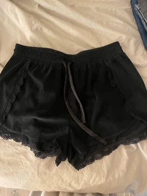 Svarta shorts med spets och snörning - Säljer ett par svarta shorts med spetsdetaljer längs kanterna och snörning i midjan. De har en loose passform och är perfekta för sommaren. Materialet känns mjukt och bekvämt, troligen bomull eller polyester. Superfina detaljer som ger en söt vibe. Köpte på Ullared 