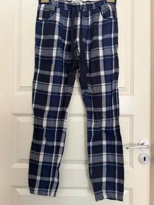 Rutiga pyjamasbyxor i blått och vitt - Mysiga pyjamasbyxor med blåvitt rutmönster och dragsko i midjan. Byxorna har två sidofickor och resår i midjan för extra komfort. Perfekta för slappa dagar hemma eller när du vill chilla i soffan.