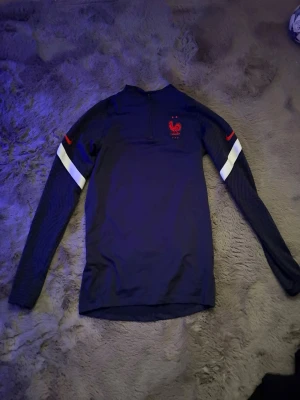 Blå långärmad träningströja Nike frankrike - Mörkblå långärmad träningströja från Nike med halv dragkedja och Frankrikes landslagsmärke på bröstet. Tröjan har vita och röda detaljer på ärmarna och är tillverkad i ett tekniskt funktionsmaterial som passar perfekt till träning.