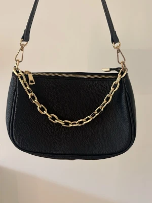 Väska - Jätte fin svart väska i läder från märket Genuine Leather. Väskan har guldfärgade detaljer och en tjock guldfärgad kedja. Väskan har också ett fack för små saker som nycklar och smink. Väskan kommer med ett långt axelband som behör till väskan.