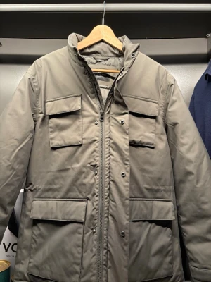 Grå Field jacket från John Henric  - Stilren grå field jacket med hög krage och fyra stora fickor framtill. Jackan har dragkedja och tryckknappar, samt en rak passform. Perfekt för kalla dagar och enkel att matcha med olika outfits. Materialet är slätt och tåligt, vilket ger en modern look. Helt ny inte använd 1 gång 