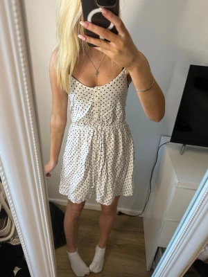Vit prickig sommarklänning - Söt vit kortklänning med svarta prickar och smala axelband. Klänningen har knappar framtill och en avslappnad passform med resår i midjan. Perfekt för varma dagar och enkel att styla med smycken eller en jeansjacka.