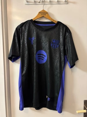 Svart Barcelona Kobe tröja - Snygg svart Barcelona matchtröja med lila detaljer och mönstrad yta. Tröjan har klubbmärke, Spotify-logga och Kobe Bryant-symbol på bröstet. Tillverkad i lätt och ventilerande material, perfekt för träning eller match. Korta ärmar och rund hals.