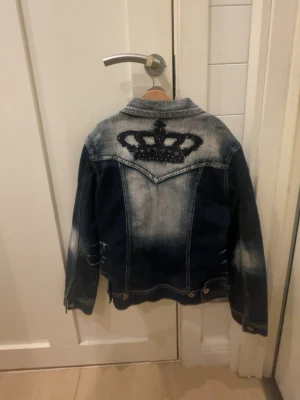 Blå jeansjacka med krona på ryggen - Snygg blå jeansjacka från crazy age med slitna detaljer och cool krona i svarta pärlor på ryggen. Klassisk modell med bröstfickor och knappstängning framtill. Perfekt för dig som vill sticka ut lite extra med din outfit.