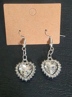 Handgjorda Hjärtformade örhängen med strass - Säljer ett par handgjorda glittriga örhängen i silverfärg med hjärtformade hängen. Örhängena har stora, klara stenar i mitten och är omgivna av små gnistrande strass. Perfekta för dig som gillar statement-smycken och vill ha något som verkligen syns.