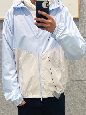 Nike ljusblå och vit vindjacka - Snygg vindjacka från Nike i ljusblått och vitt med dragkedja framtill och huva. Jackan har två sidofickor, elastiska muddar och en sportig design med färgblock. Perfekt för dig som gillar streetwear och vill ha en lätt jacka till vardags.