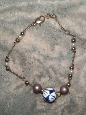 Armband med pärlor och blå detalj - Unikt armband med små vita och silverfärgade pärlor på tunn guldfärgad kedja. I mitten sitter en större blåvit pärla med handmålat mönster och två lila pärlor på varsin sida. Perfekt accessoar för dig som gillar personliga smycken.