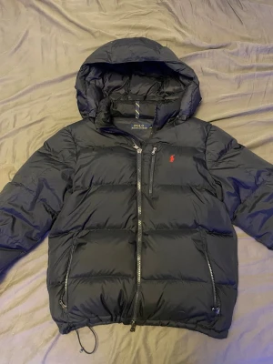Marinblå pufferjacka från Polo Ralph Lauren - Marinblå pufferjacka med huva från Polo Ralph Lauren. Jackan har dragkedja framtill, två sidofickor med dragkedja och en bröstficka. Klassisk röd logga broderad på bröstet. Vadderad och perfekt för kalla vinterdagar.