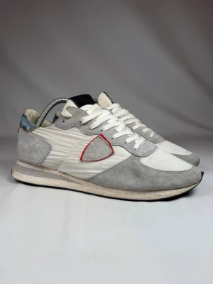 Philippe Model vita/grå sneakers - Storlek 43 men passar för 44. Snygga sneakers från Philippe Model i vitt och ljusgrått med mockadetaljer och mesh. Skon har en ikonisk sköldlogga med röd kant på sidan, vita skosnören och en retroinspirerad sula. Perfekt för dig som gillar stilrena och sportiga vibes.
