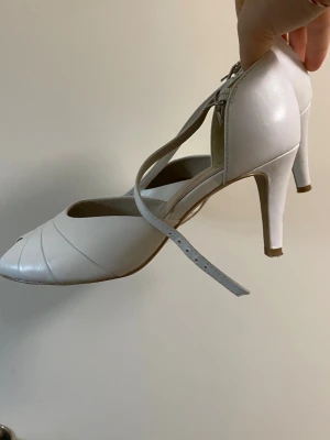 Vita vintage peep toe pumps från Caprice - Säljer ett par vita vintage pumps från Caprice med öppen tå och smala ankelremmar med spänne. Skorna har en klassisk klack och är tillverkade i ett slätt, glansigt material som ger en elegant look. Perfekta för dig som vill ha stilrena och feminina skor.