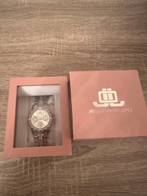Silverfärgad klocka från JLO med stenar - Snygg silverfärgad klocka från Jennifer Lopez med glittrande stenar runt urtavlan och på armbandet. Klockan har en rund urtavla med flera små visare och ett elegant metallarmband. Perfekt accessoar för att lyfta din stil. Dock fungerar inte klockan därav priset men det går ävan att diskuteras vid snabb affär! OBS: FRAKTAR INOM 24H!🩷