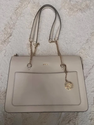 Beige handväska från DKNY med kedjedetaljer - Snygg beige handväska från DKNY i stilren, rektangulär modell. Väskan har guldfärgade kedjor och detaljer, samt ett dekorativt DKNY-mynt. Utsidan är i slätt konstläder med en stor ficka framtill och dubbla axelremmar. Perfekt för dig som gillar klassisk och trendig design.